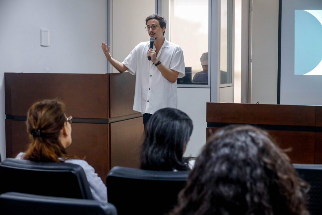 Professor Túlio Rodrigues fala em microfone para turma de alunos, que estão de costas. Ele é um homem magro, alto, usa óculos e veste uma camisa branca com botões escuros. O local é um auditório com paredes brancas e móveis em madeira escura. É possível ver a parte de uma tela com a projeção do conteúdo da aula.